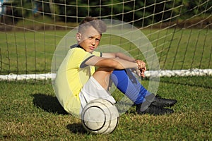 Boy football player in a yellow t-shirt sits near the goal with the ball
