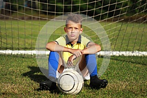 Boy football player in a yellow t-shirt sits near the goal with the ball