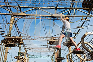 Boy in forest adventure park. Kid in orange helmet and white t shirt climbs on high rope trail. Climbing outdoor