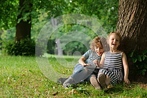 Boy with girl of 7-8 years sit under a tree having nestled and play on the tablet.