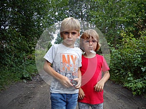 A boy and a girl are brother and sister. Children with blond hair walk next to trees and bushes. Girl with glasses and a red t-