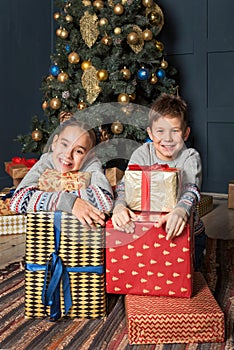 Boy and girl, brother with sister, siblings are sitting near the Christmas tree smiling happily hugging gifts boxes