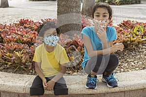 Boy and girl casually dressed sitting with face masks outdoor on a retaining wall