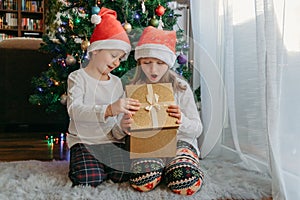 A boy and a girl in Christmas hats open a gift box under a Christmas tree. Celebration, receiving gifts, children`s joy. Miracle,