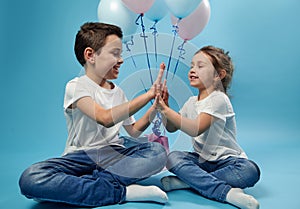 Boy and girl clapping hands while posing on blue background with balloons behind them