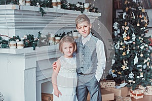 Boy and girl dressed elegantly standing in a bright room by the fireplace. Christmas tree in the background. New year concept