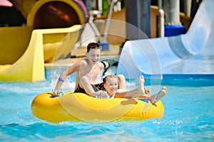 Boy and girl have fun on water slide in outdoor aquapark. Little children floating on yellow inflatable raft.