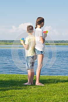Boy and girl on the lake, holding water pistols and stand with their backs to each other, against a beautiful landscape, looking