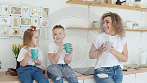 Boy, girl and mother joke and drink a drink from blue mugs sitting on the table in a stylish white modern kitchen in