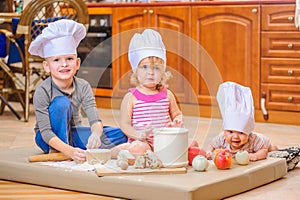 Boy and girl and a newborn kid with them in chef`s hats sitting on the kitchen floor soiled with flour