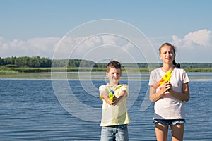 Boy and girl play water pistols against the blue lake and sky