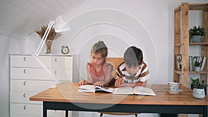 A boy and a girl are reading a book sitting on a table. The children are doing their homework. Home education