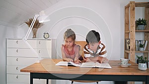 A boy and a girl are reading a book sitting on a table. The children are doing their homework. Home education