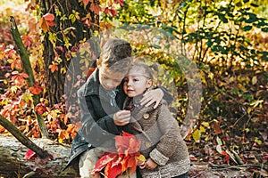 Boy and girl with red leaves in hands sitting and embracing together on the log in the autumn park