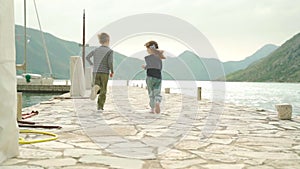 A boy and a girl run along the pier against the backdrop of the sea and mountains