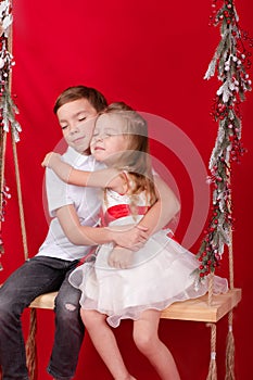 boy and girl - sister and brother - sitting on a swing decorated with christmas decor and tree branches. on red