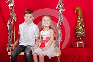 boy and girl - sister and brother - sitting on a swing decorated with christmas decor and tree branches. on red
