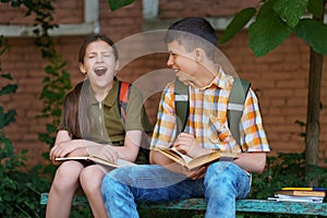 A boy and a girl are sitting on a bench near the school building, they are reading books, the girl is bored, she yawns and wants