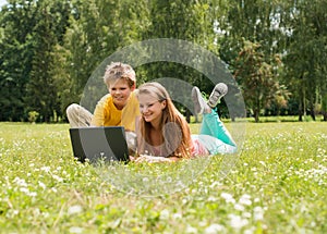 Boy and girl sitting on grass with laptop, online in park. Two smiling teenagers students with laptop resting on meadow. Education