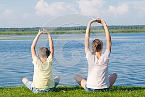 A boy and a girl are sitting on the green grass and doing yoga, holding their hands above their heads, rear view, against the