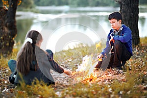 Boy and girl sitting on the river Bank, make a fire, autumn forest at sunset, beautiful nature and reflection of trees in the wate