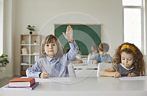 Boy and a girl are sitting at a school desk, writing, the boy raises his hand up.