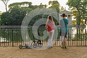 Boy and girl with their push scooters against the backdrop of the Tempio di Esculapio in the park Villa Borghese