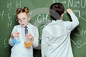 Boy and girl in white coats standing with reagents in flasks and drawing chemical formulas