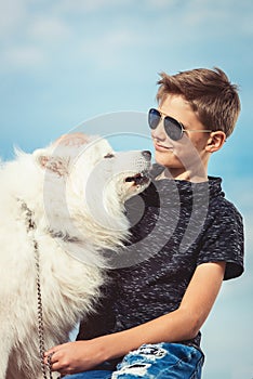 Happy 11 year old boy playing with his dog breed Samoyed at the seashore against a blue sky close up. Best friends rest