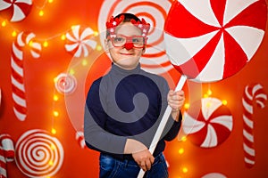 A boy in glasses with reindeer horns and a red nose holds in his hands a large toy candy