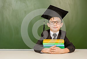 Boy in graduation cap with books sitting near green chalkboard