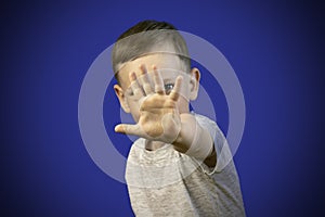 A boy in a gray T-shirt put his hand forward and covered his face with his palm. The child hid behind his hand. Stop gesture. Blue