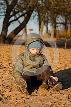 A boy in a green jacket, a blue knitted hat, brown trousers and boots stands on the yellow sand and looks down, stained his pants