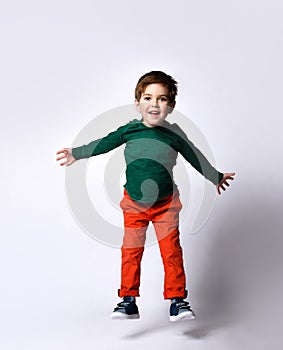 Boy in green jumper, orange pants, blue sneakers. He jumping with raised hands, smiling, posing isolated on white