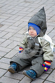 A boy in green snowsuit sitting on paving stone