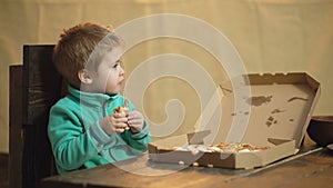 Boy in a green sweater eats a pizza while sitting on a wooden chair. Tasty pizza. Little boy holds slice of pizza