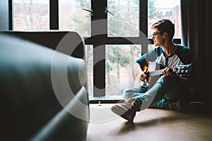 Boy with guitar sits on the floor at cozy home, moody day light