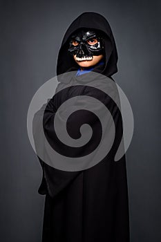 A boy in a Halloween costume dressed in a black cloak with a hood and a black skull mask s