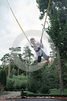 Boy hangs on slings, jumps high on trampoline in an amusement park. Weekend at theme park. Teenager is having fun