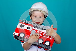 A boy in a hard hat holds a toy fire truck in his hands and smiles and shows a thumbs up. Isolated on a blue background