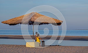 A boy in a hat and a girl on the beach with a suitcase under a straw umbrella look into the distance. Traveling of children