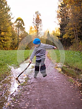 Boy in hat running along the path and strikes stick on puddle against the sky