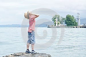 Boy in hat standing on the beach and looking up. Small lighthouse in the distance. Back view