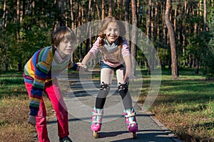 Boy helps the girl to roller-skate in the park. Brother supports