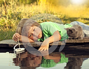 Boy and his beloved kitten playing with a boat from pier in pond