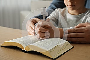 Boy and his godparent praying together at wooden table indoors, closeup