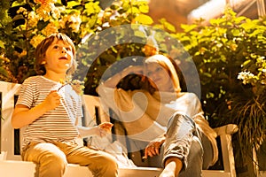 Boy and his mom sitting on the bench in city park. Son and mother having fun on log seat with bushes in the background