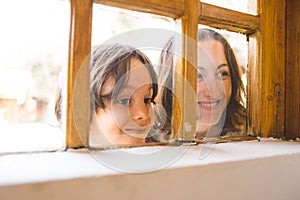 A boy with his mother curiously peeks out the window of a wooden house