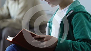 Boy holding book learning to read with father sitting on sofa, lesson with tutor