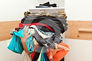 Boy holding a huge pile of clothes. Kid makes order in the closet. Storage organization